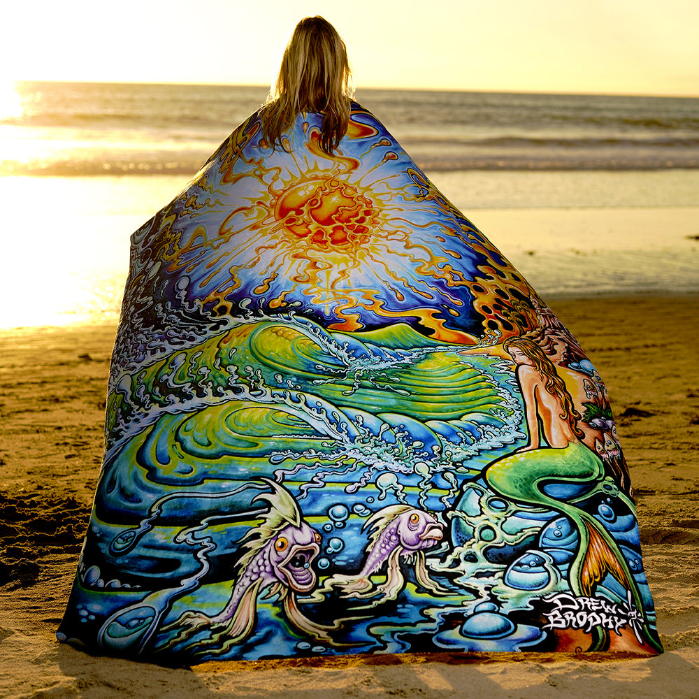 Person wrapped in a colorful "Mystic Sun" blanket by Drew Brophy with mermaid design on a beach at sunset