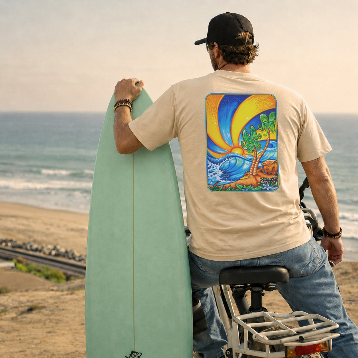 Surfer's Paradise Tee by Drew Brophy - A man on a beach stands with a surfboard, wearing a beige t-shirt with a colorful graphic print on the back, blue jeans, and a black cap. He is positioned next to a bicycle with the ocean and sandy beach in the background.